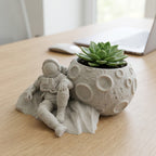 CONQR astronaut moon planter with a green succulent on a light oak office desk beside a laptop