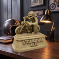 Still Raising Hell XL (129 mm) on a dark desk, shown to scale beside a motorcycle helmet and lamp